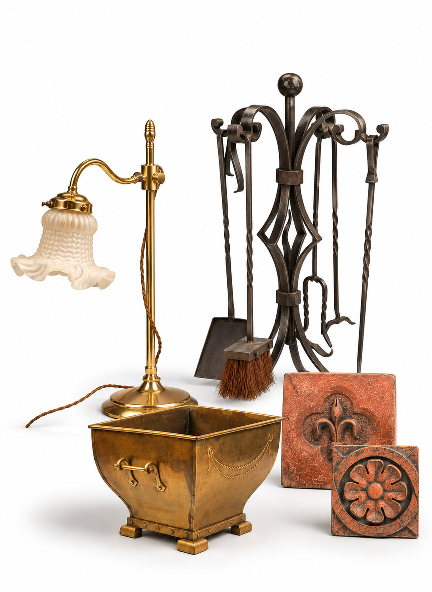 Curated selection of antique lighting, fireside accessories and decorative antiques from the Shackleton Fox Sale Collection, displayed against a clean white background. The arrangement includes an elegant brass table lamp with a curved arm and decorative frosted glass shade, a dark wrought iron fireside companion set with scrolling metalwork, a rectangular brass coal scuttle with decorative handles and feet, and two terracotta-coloured decorative plaques featuring raised floral and fleur-de-lis motifs.