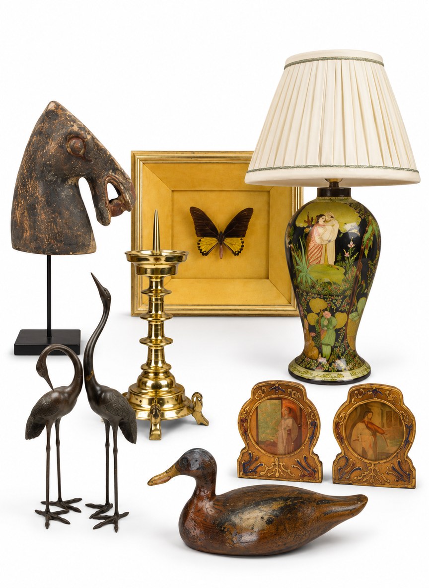 Curated display of sold antiques and decorative objects from Shackleton Fox, including an antique carved wooden horse head on a black display stand, a polished brass ecclesiastical candlestick, two bronze crane sculptures, a colourful hand-painted ceramic table lamp with a cream pleated shade, a framed butterfly specimen, two small gilt decorative picture frames and a carved wooden duck decoy. The collection is photographed against a clean white background, showcasing the variety of antique lighting, artwork, folk art and decorative antiques previously sold by Shackleton Fox.